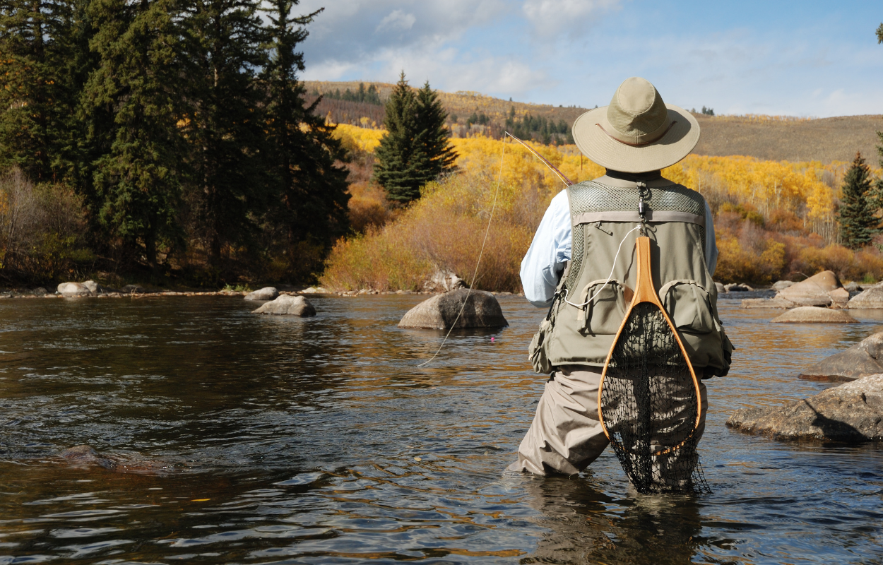 fly fishermen stands in a Colorado trout stream with a vest on and a net hanging off his back