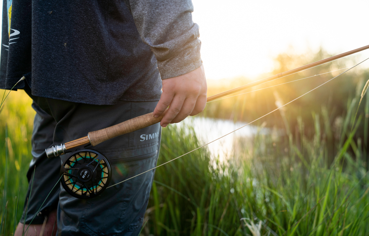 man stands with bamboo fly fishing rod at his side on a grassy stream at sunset