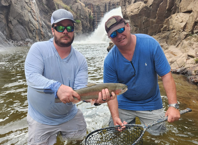 two men hold rainbow trout at the base of a waterfall