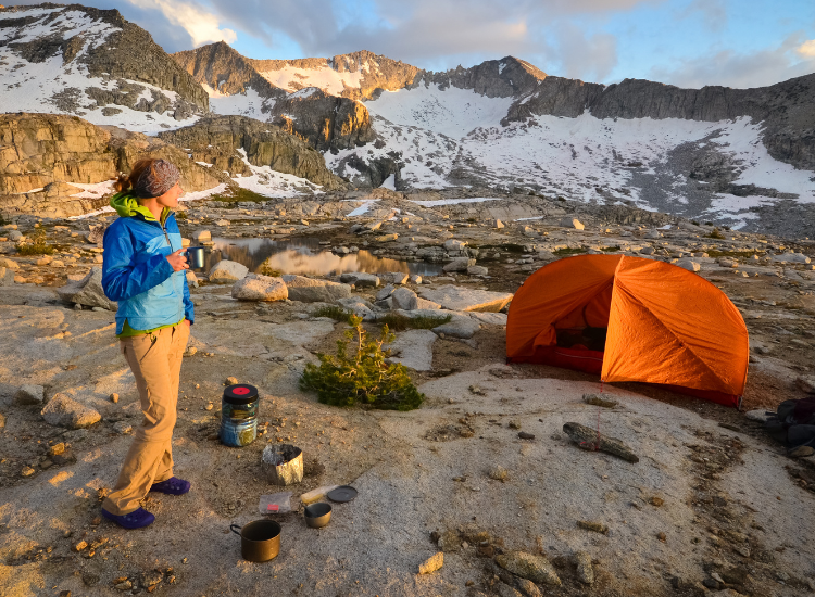 a backpacker stands in the mountains next to an orange tent